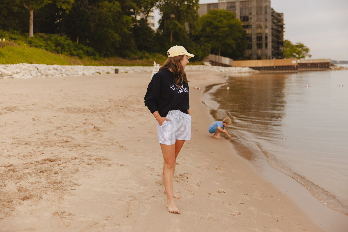 Jen in the Third Coast crewneck and yellow cap, walking the shoreline while her son plays at the water